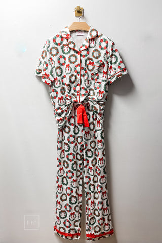 Jess Lea Ribbon Wreath Ruffle Pajama Set on a hanger, ivory short sleeve button-down top and matching pants with a Christmas wreath and red bow print, red piping, red tie detail, and red ruffle hem