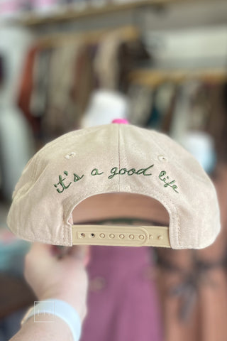 Back of tan baseball cap with "it's a good life" embroidered in green script above an adjustable snapback closure