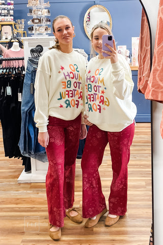 Two models wearing the Poppy & Pine So Much To Be Grateful For ivory oversized graphic sweatshirt with multicolor lettering, styled with red flare jeans, size S/M